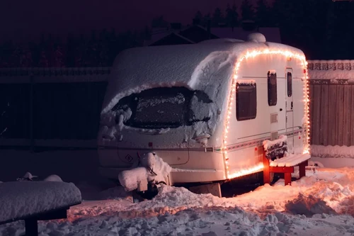 A snow-covered RV with holiday lights glowing at night, showing heavy winter buildup on the roof and windows. 